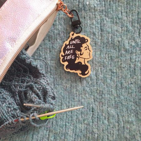a wooden charm, depicting a long-haired person in black ink. on their hair is printed: "until all are free". the charm is attached to a vinyl case holding a piece of half-finished knitting.